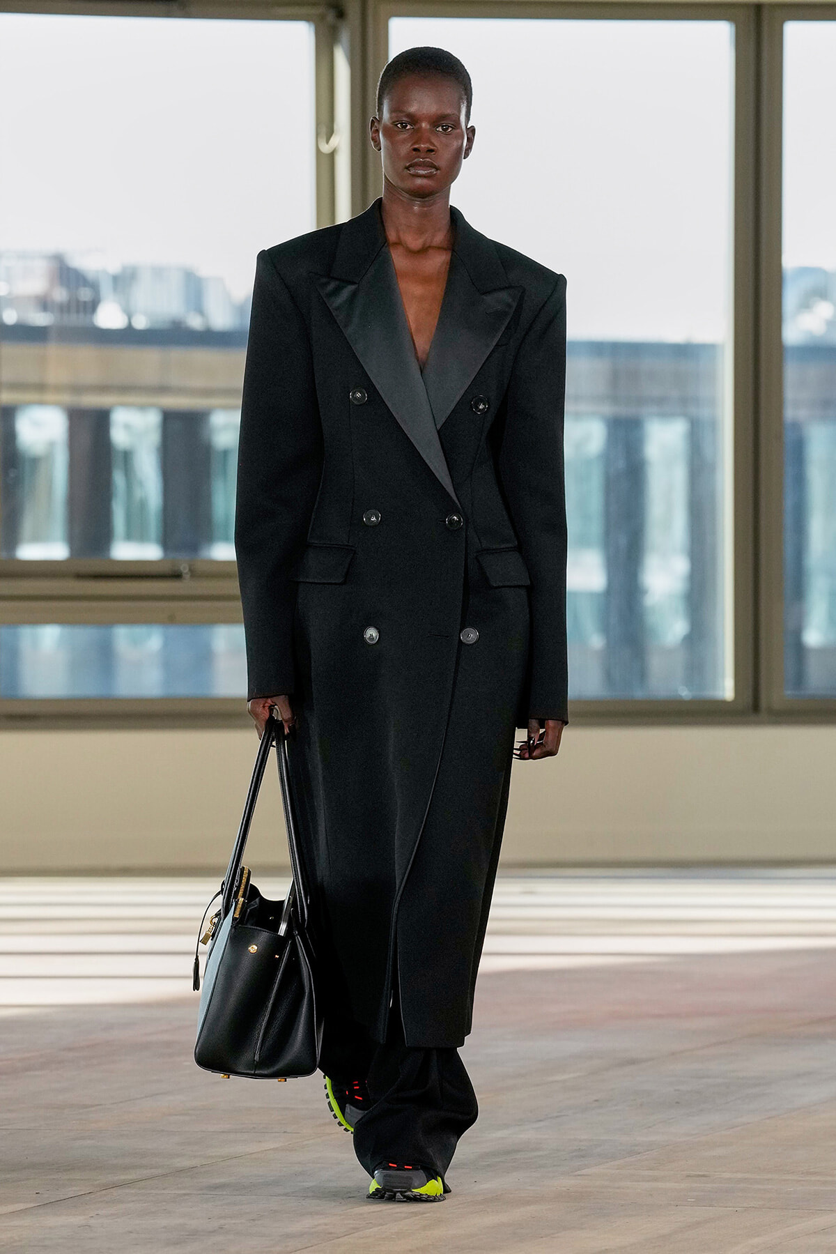 Model in a long black double-breasted coat, holding a black handbag, walking in a bright studio with large windows.