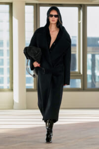 Woman in an oversized black coat with hood and sunglasses, carrying a black braided bag, walking indoors in a bright modern space.