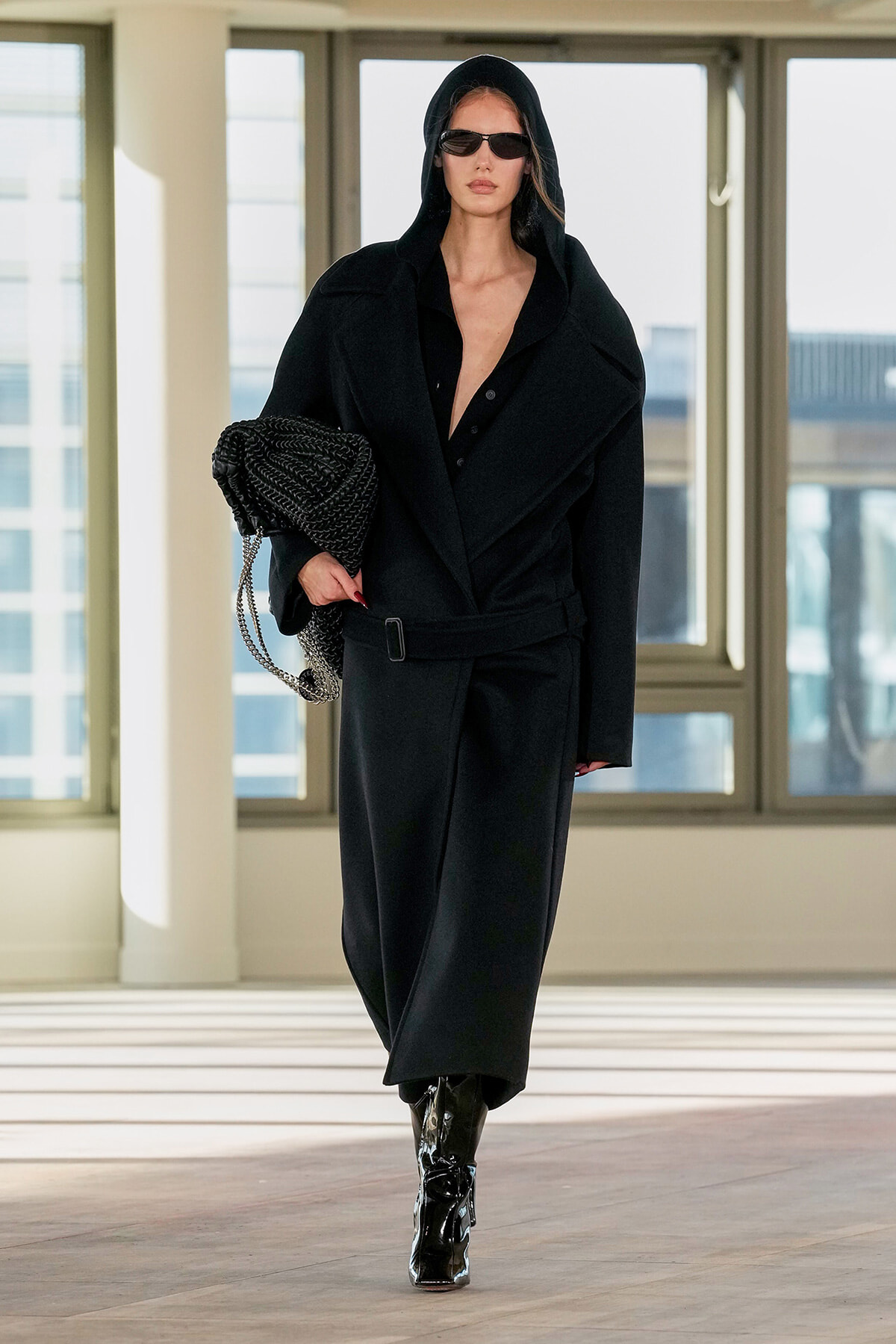 Woman in an oversized black coat with hood and sunglasses, carrying a black braided bag, walking indoors in a bright modern space.
