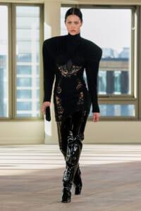 Model walks in a black ensemble with lace panels and shiny thigh-high boots in a bright studio setting