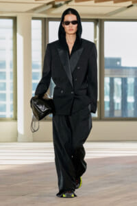 Model wearing a black oversized double-breasted blazer with a hood and sunglasses, carrying a glossy black clutch indoors.