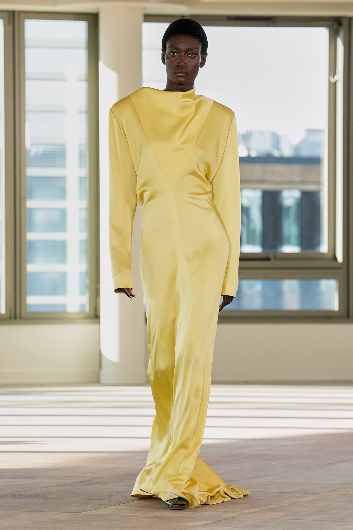 Model wearing a pale yellow satin, long-sleeve, floor-length gown with subtle seam details, standing in a bright studio.