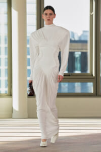 Model wearing a white sculptural gown with a heart-shaped bodice, ruched sides, and long sleeves, carrying a small dark handbag indoors.