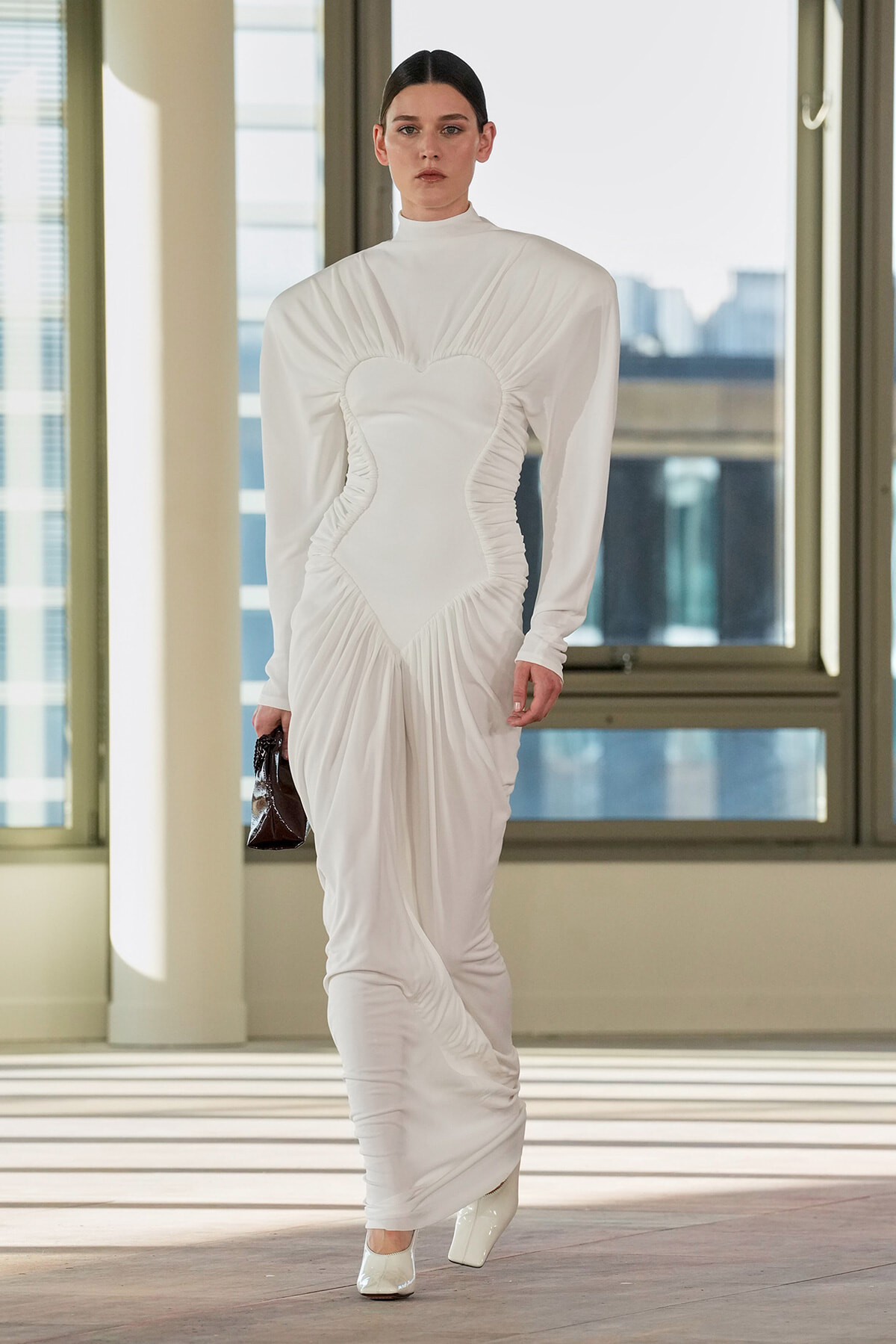 Model wearing a white sculptural gown with a heart-shaped bodice, ruched sides, and long sleeves, carrying a small dark handbag indoors.