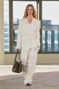 Model walks in a white double-breasted suit, carrying a dark brown handbag on a runway.