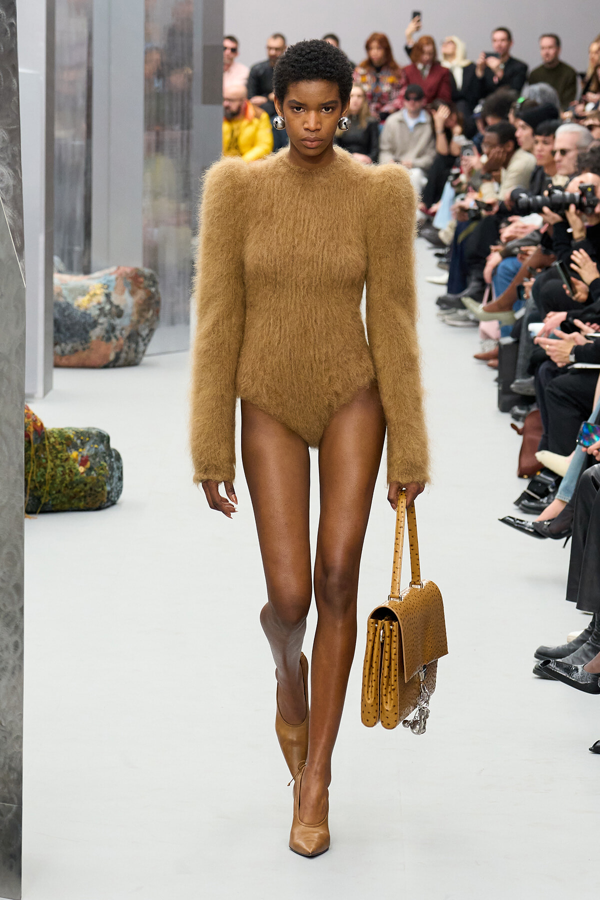 Runway model in a fuzzy tan bodysuit with padded shoulders, tan heels, and a matching tan handbag as photographers look on.