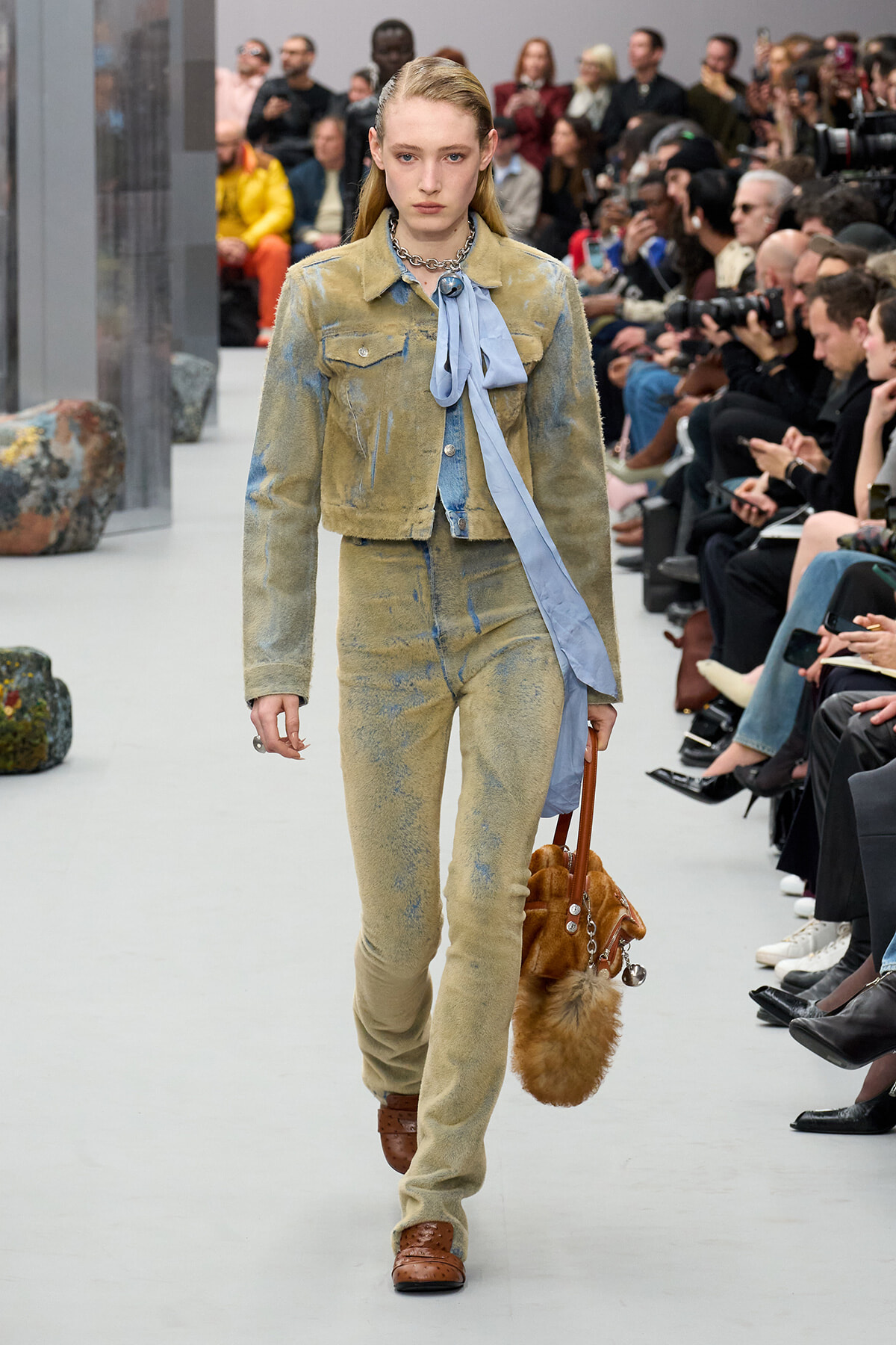 Model walking a fashion show in a distressed beige denim suit with a light blue scarf hanging from the neck, audience in background.