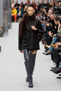 Model walks the runway in a black coat with a plush fur collar and tall gray studded boots, audience watching in the background.