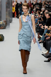 Model walking a fashion runway in a light blue ruffled dress with brown knee-high boots and a dark necklace as photographers watch from the sides.