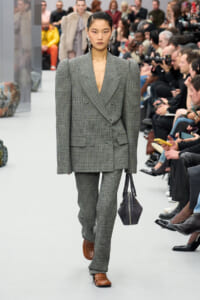 Runway model wearing a gray check tailored double-breasted suit with broad shoulders, matching trousers, brown loafers, and a dark handbag, walking the catwalk.