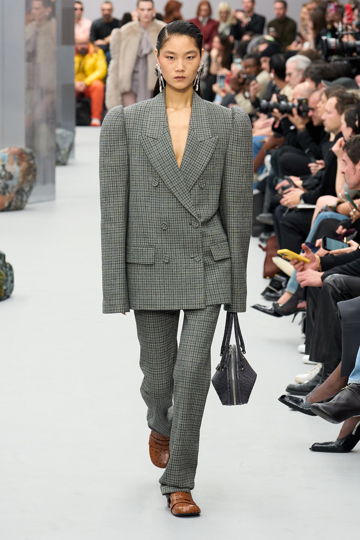 Runway model wearing a gray check tailored double-breasted suit with broad shoulders, matching trousers, brown loafers, and a dark handbag, walking the catwalk.