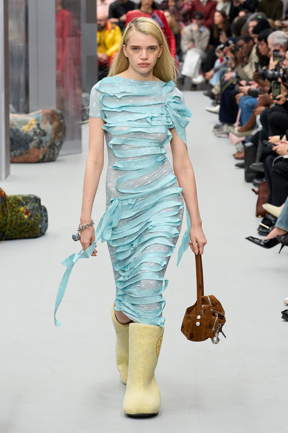 Runway model walking in a sky-blue ruched dress with ribbon-like layers and cream-yellow boots, carrying a brown handbag.