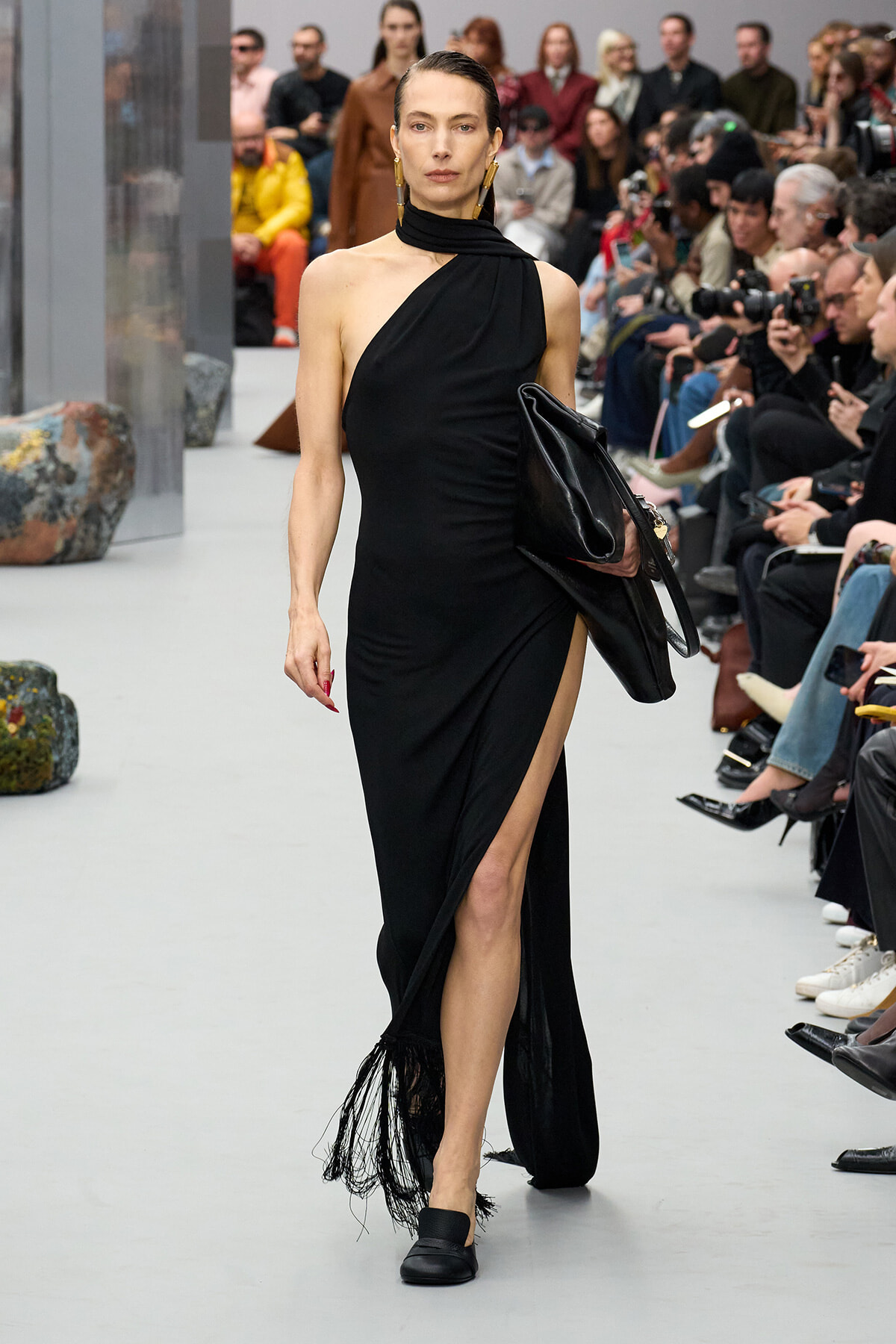 Model walking a fashion runway in a black one-shoulder gown with a high slit, carrying a large black clutch with fringe at the hem