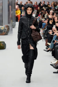 Runway model in a black hooded jacket and tall boots, carrying a brown bag, walking past seated spectators.
