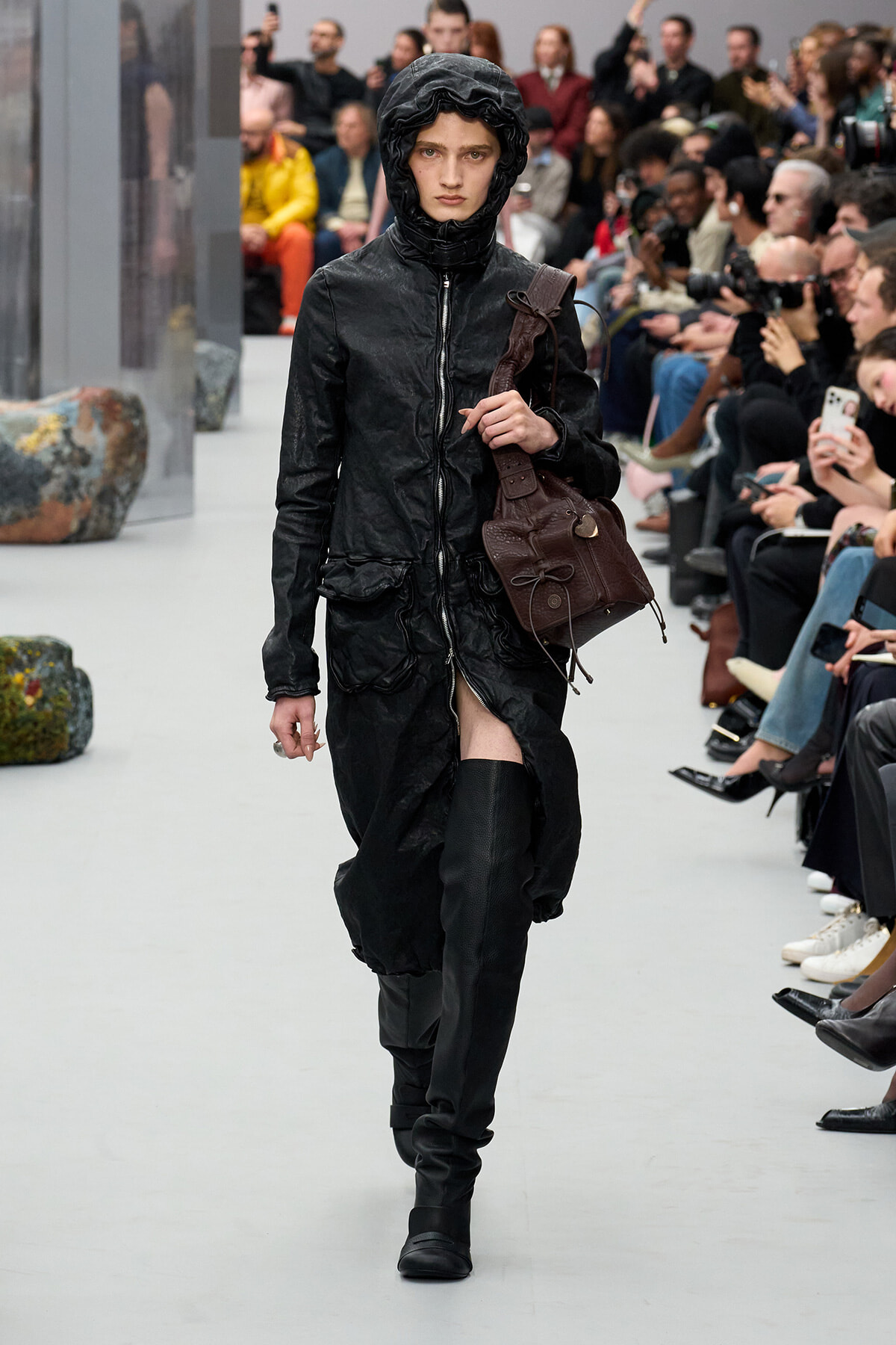 Runway model in a black hooded jacket and tall boots, carrying a brown bag, walking past seated spectators.
