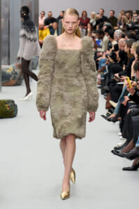 Model walks a fashion runway in a plush, gray, knee-length dress with oversized puff sleeves and a square neckline; gold pointed-toe heels and a seated audience frame the scene.
