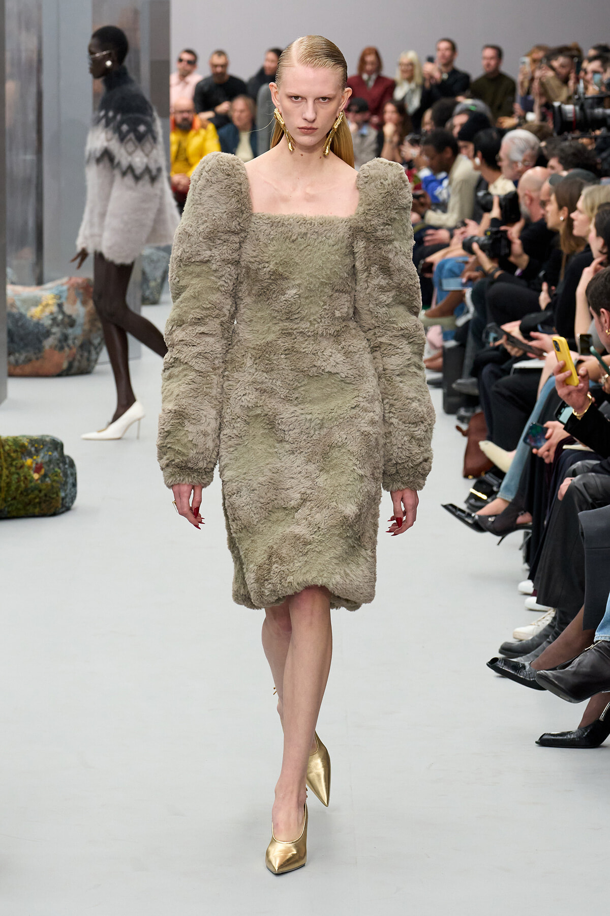 Model walks a fashion runway in a plush, gray, knee-length dress with oversized puff sleeves and a square neckline; gold pointed-toe heels and a seated audience frame the scene.
