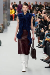 Model on a runway wearing a navy textured bow-front top, burgundy midi skirt, white knee-high boots, and a brown handbag. Audience watches in the background.