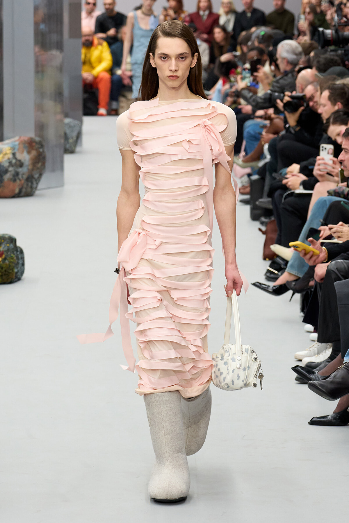 Model walking a fashion runway in a pale pink, ribbon-ruffled dress, paired with light gray knee-high boots and a white star-pattern handbag as photographers watch.