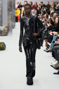 Model walks a runway in a glossy black leather jumpsuit with bold jewelry, carrying a black bag as the audience watches.