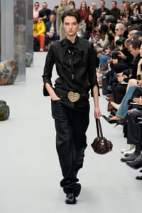 Model walks the runway in an all-black leather ensemble with a tied blouse, large ornate heart belt buckle, and a studded brown handbag; spectators line the catwalk.