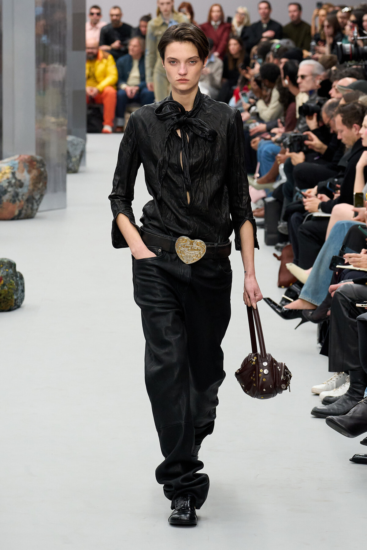 Model walks the runway in an all-black leather ensemble with a tied blouse, large ornate heart belt buckle, and a studded brown handbag; spectators line the catwalk.
