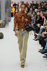 Model walks a runway in a brown leather blouse with a bow, a wide heart-buckle belt, paint-splattered beige jeans, brown boots, and a black handbag as the audience watches.