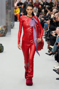 Model walking a fashion show wearing a red leather jumpsuit with a lavender scarf tied at the neck and a red bag, audience in background.