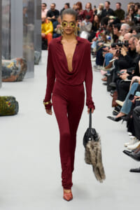 Model walks the runway in a burgundy draped jumpsuit with a deep neckline, gold earrings, sunglasses, orange heels, and a fur-trim handbag.