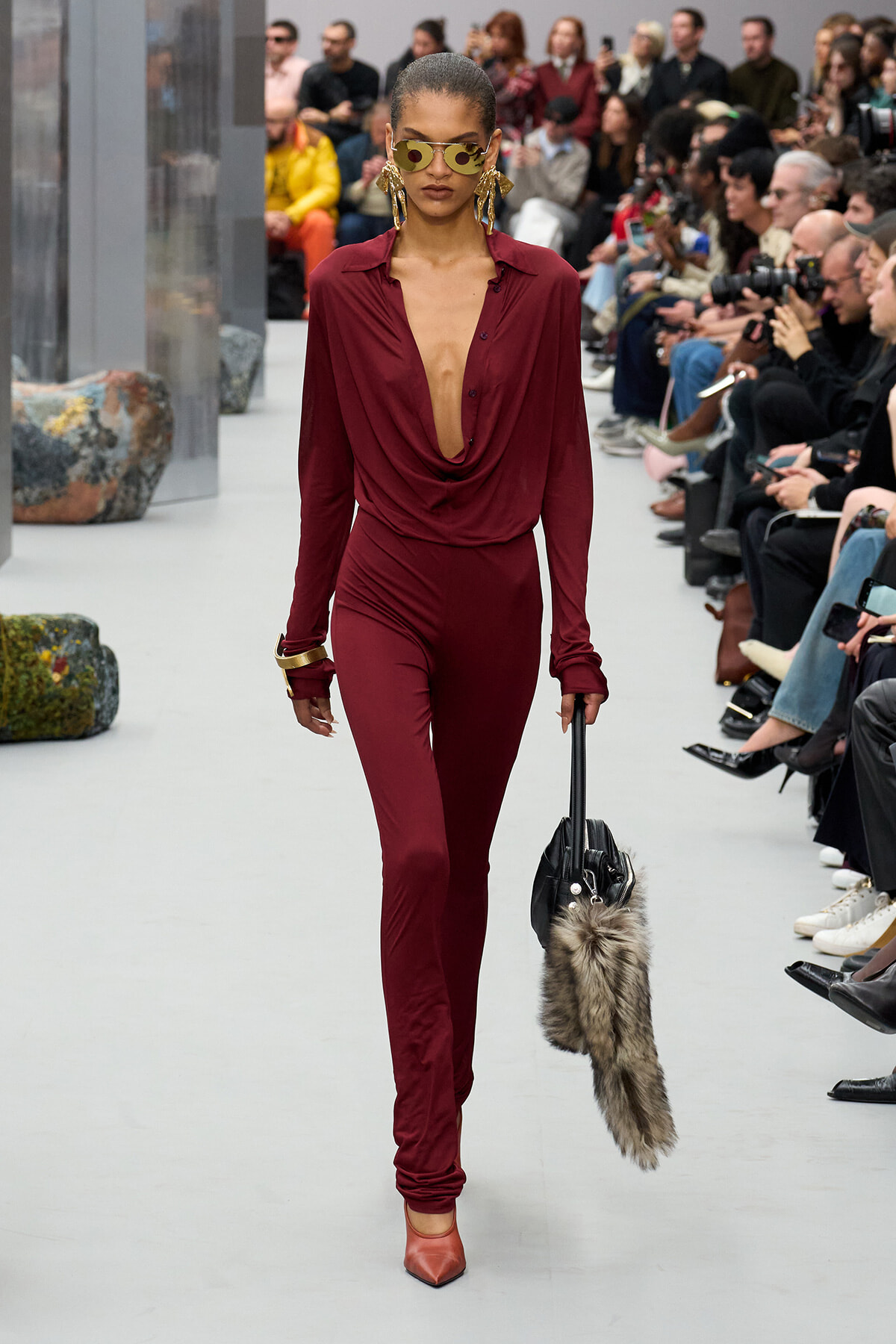 Model walks the runway in a burgundy draped jumpsuit with a deep neckline, gold earrings, sunglasses, orange heels, and a fur-trim handbag.