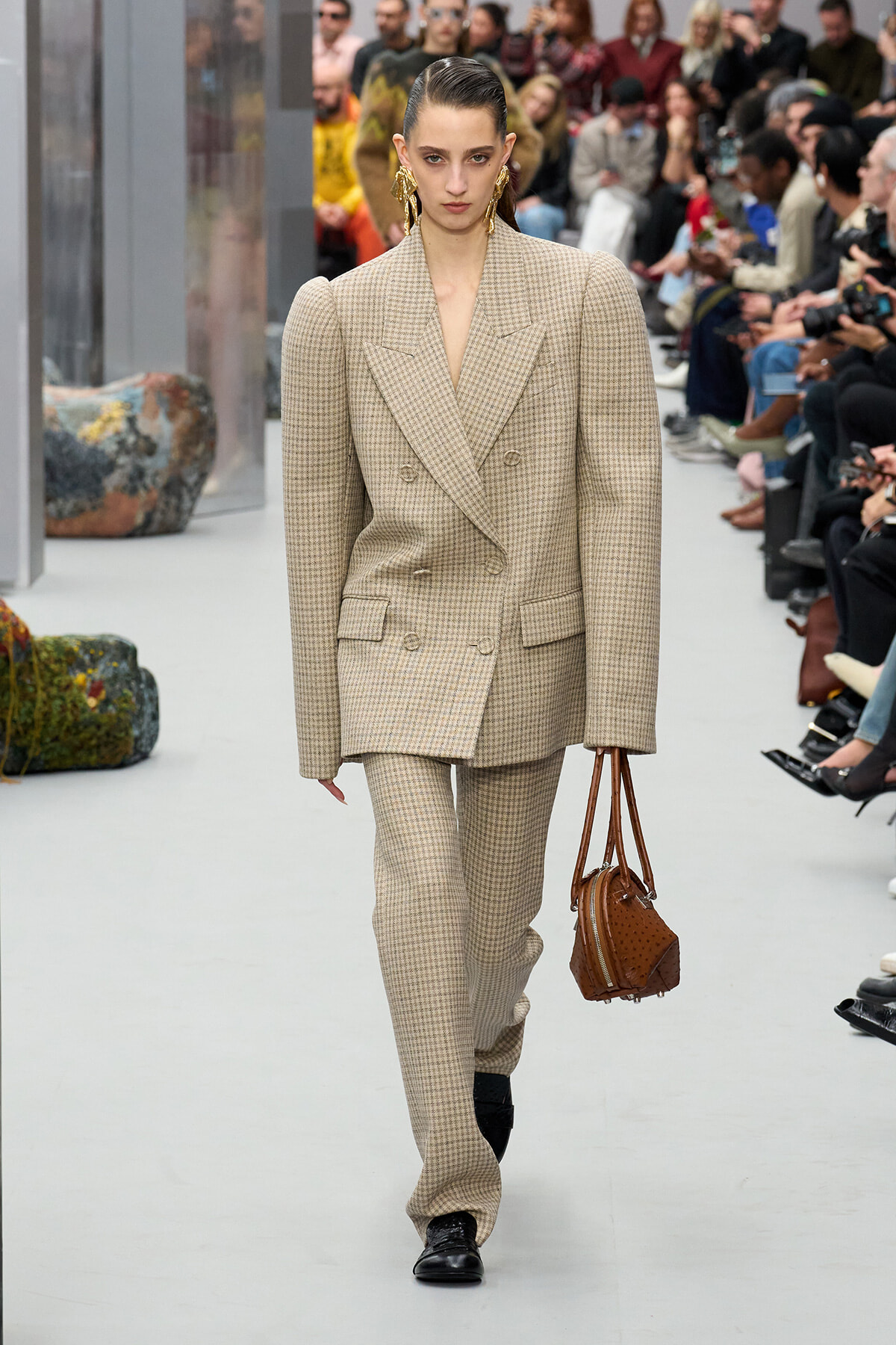 Female model walks a runway in a beige checked oversized double-breasted suit, carrying a brown handbag; audience seated in the background.