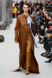 Model walks the runway in a brown leather trench with oversized buttons, a blue scarf, mustard yellow polka-dot boots, and a dark brown bag.