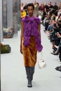 Model walks a runway in a bold purple textured top tied at the neck, with a brown faux‑fur midi skirt and gray knee‑high boots, carrying a white handbag.