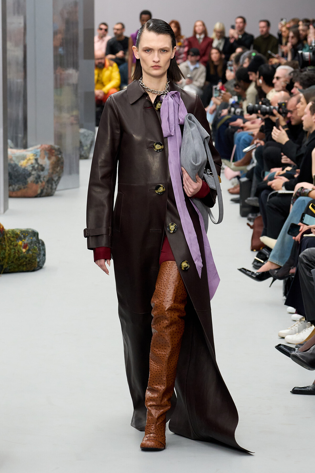 Model walking the runway in a dark leather coat with a purple scarf and burgundy outfit, paired with tan perforated boots.