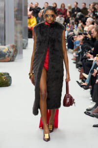Model walks a runway in a black fur vest over a red outfit, wearing sunglasses and gold-buckled burgundy heels, carrying a small burgundy handbag.