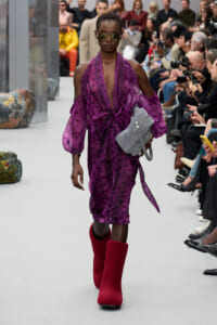 Model walks the runway in a purple star-patterned dress with cold-shoulder sleeves, red knee-high boots, sunglasses, and a gray handbag peacefully tucked under her arm.