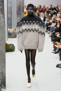 Model on a runway wearing an oversized fuzzy gray-to-white knit sweater and white heels, audience in background.