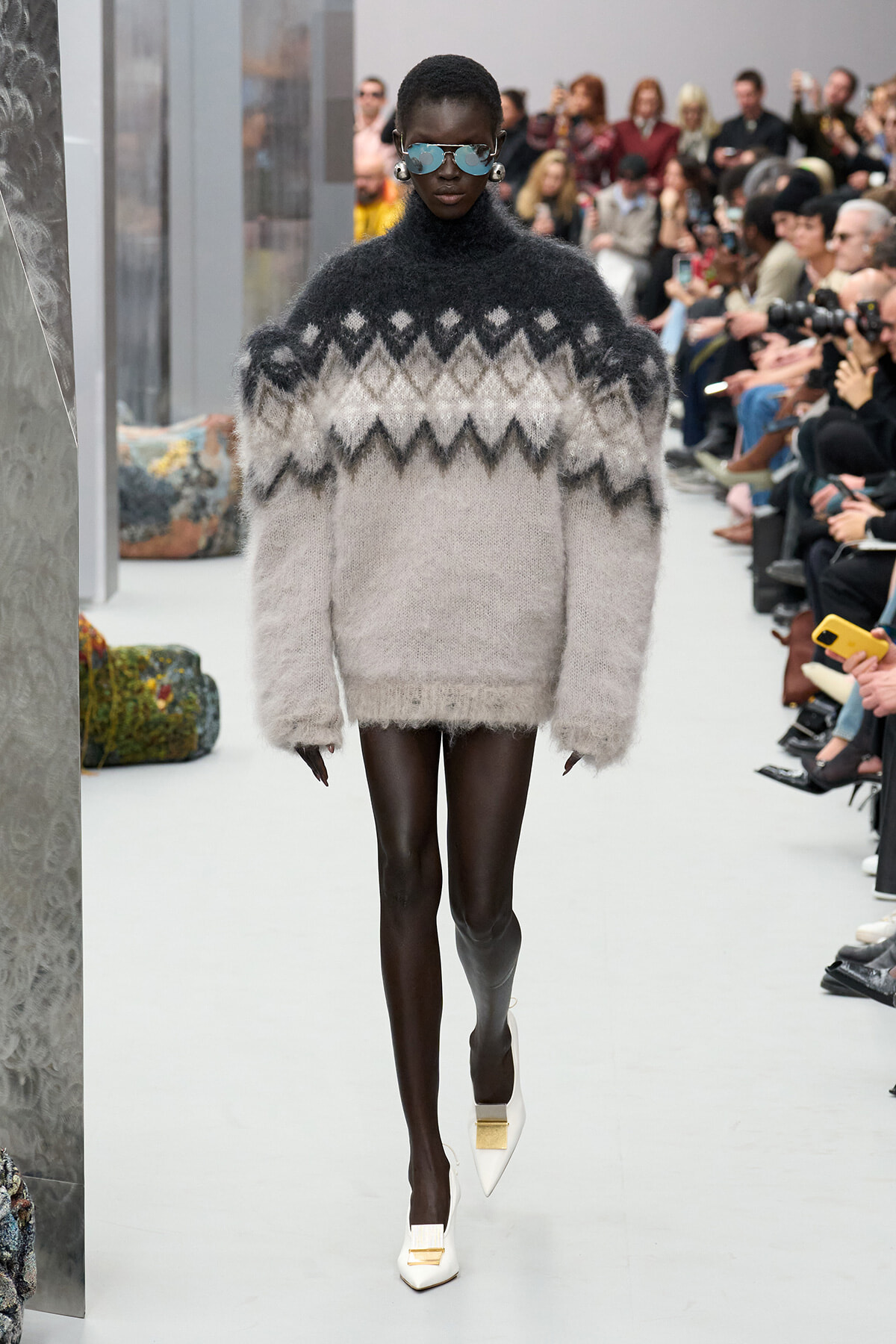 Model on a runway wearing an oversized fuzzy gray-to-white knit sweater and white heels, audience in background.