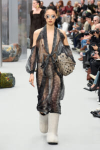 Model walks a fashion runway in a sheer gray star-print dress with a waist tie, oversized sleeves, white knee-high boots, sunglasses, and a star-cutout handbag; audience in background.