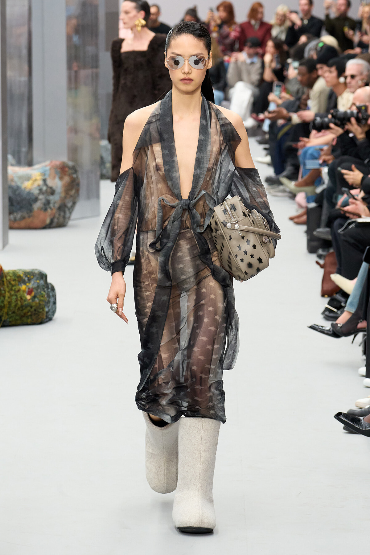 Model walks a fashion runway in a sheer gray star-print dress with a waist tie, oversized sleeves, white knee-high boots, sunglasses, and a star-cutout handbag; audience in background.
