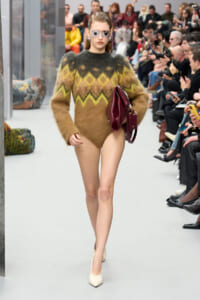 Model walking a runway in a fuzzy multicolor zigzag knit sweater, carrying a burgundy handbag and wearing reflective sunglasses.