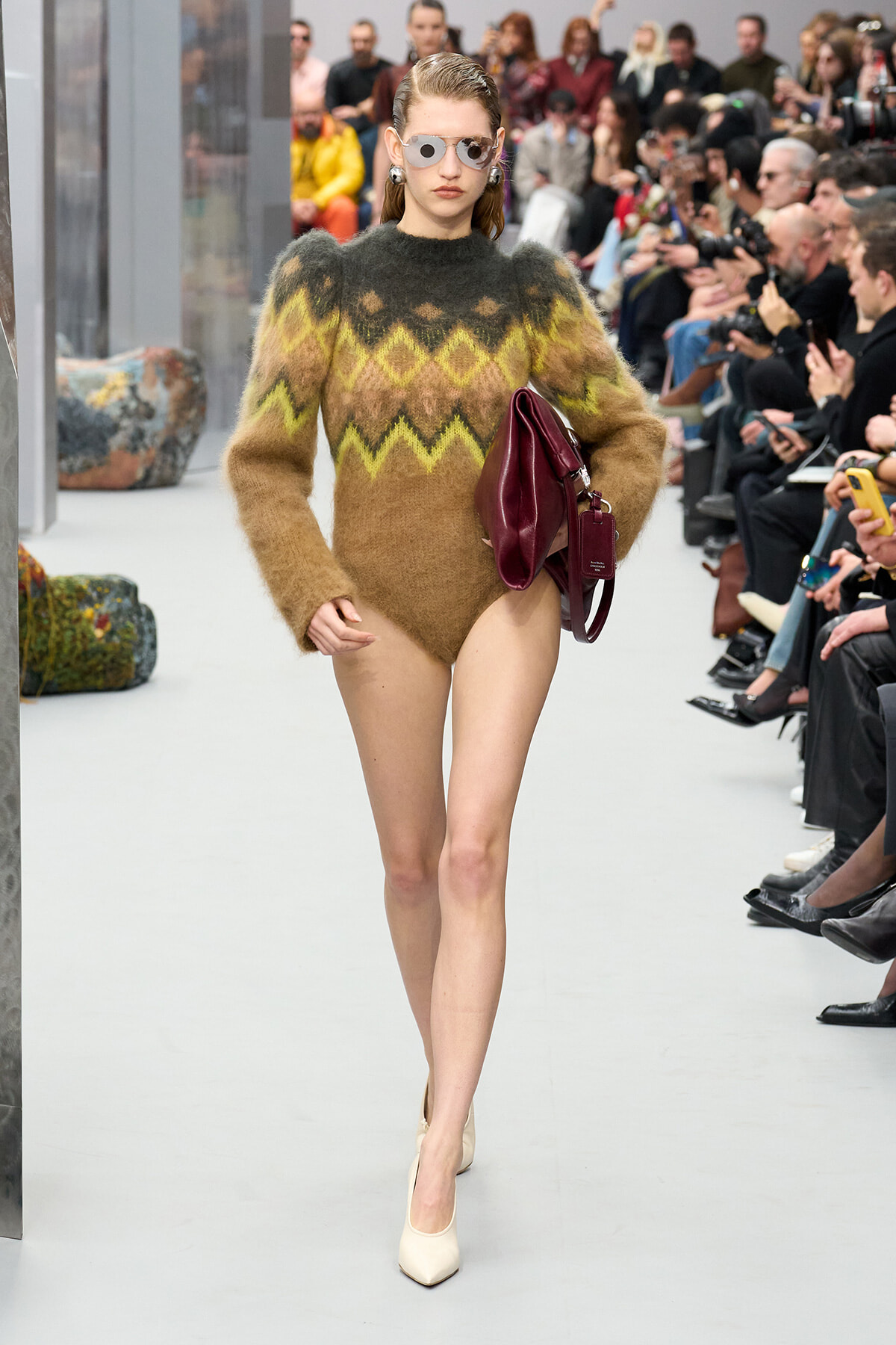 Model walking a runway in a fuzzy multicolor zigzag knit sweater, carrying a burgundy handbag and wearing reflective sunglasses.
