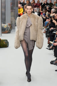 Model on a fashion runway wearing an oversized beige fur coat over a gray blouse and draped skirt, black tights, and dark pointed heels as spectators watch.