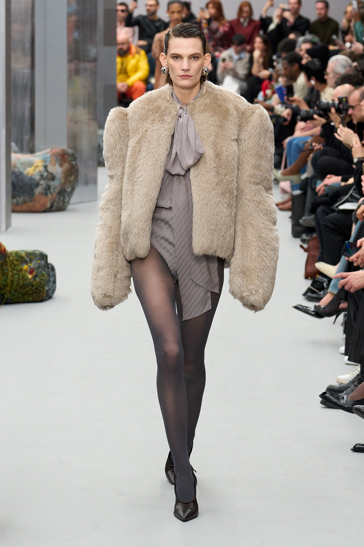 Model on a fashion runway wearing an oversized beige fur coat over a gray blouse and draped skirt, black tights, and dark pointed heels as spectators watch.