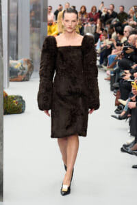 Model walks a fashion show in a knee-length black textured fur dress with long sleeves and off-the-shoulder neckline, paired with gold earrings and black heels with a white-gold buckle.