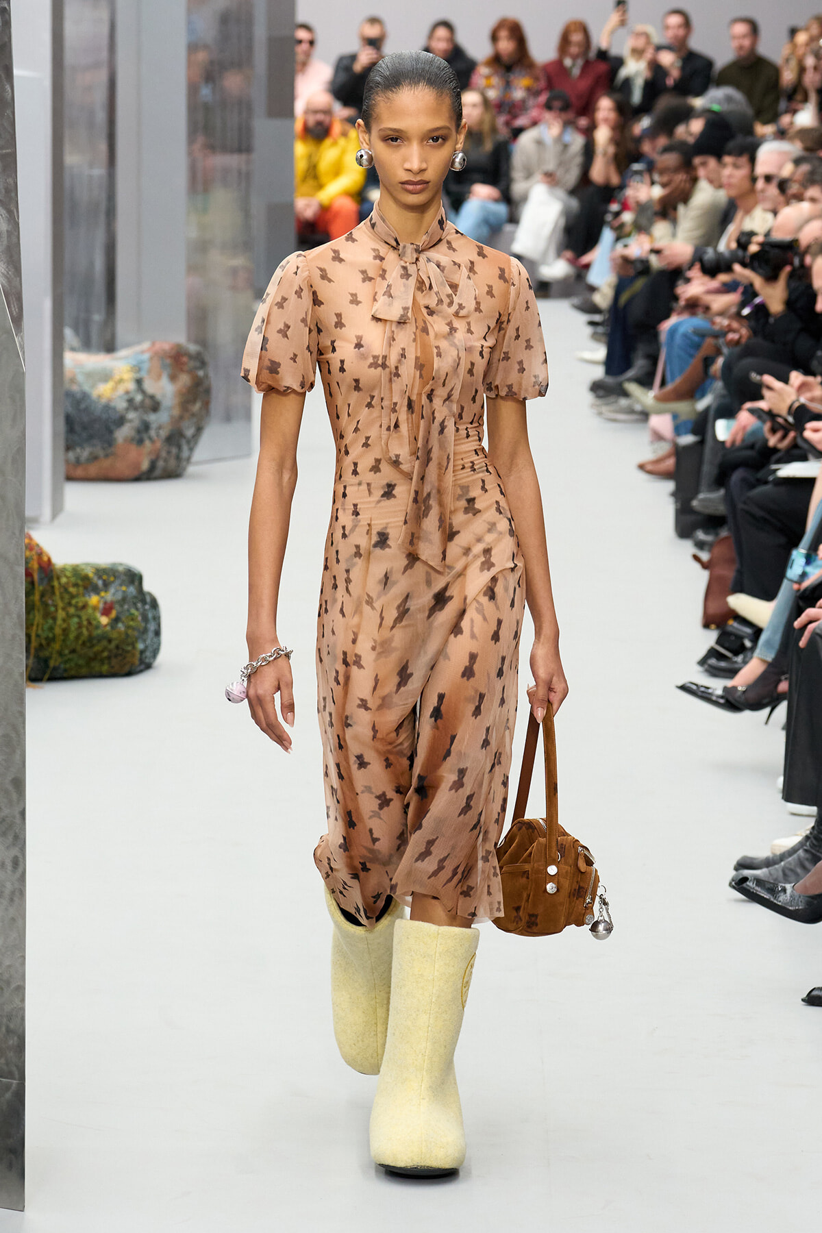 Model walking a fashion runway in a peach-toned floral dress with a neck bow, carrying a brown handbag and wearing yellow knee-high boots, audience in the background.