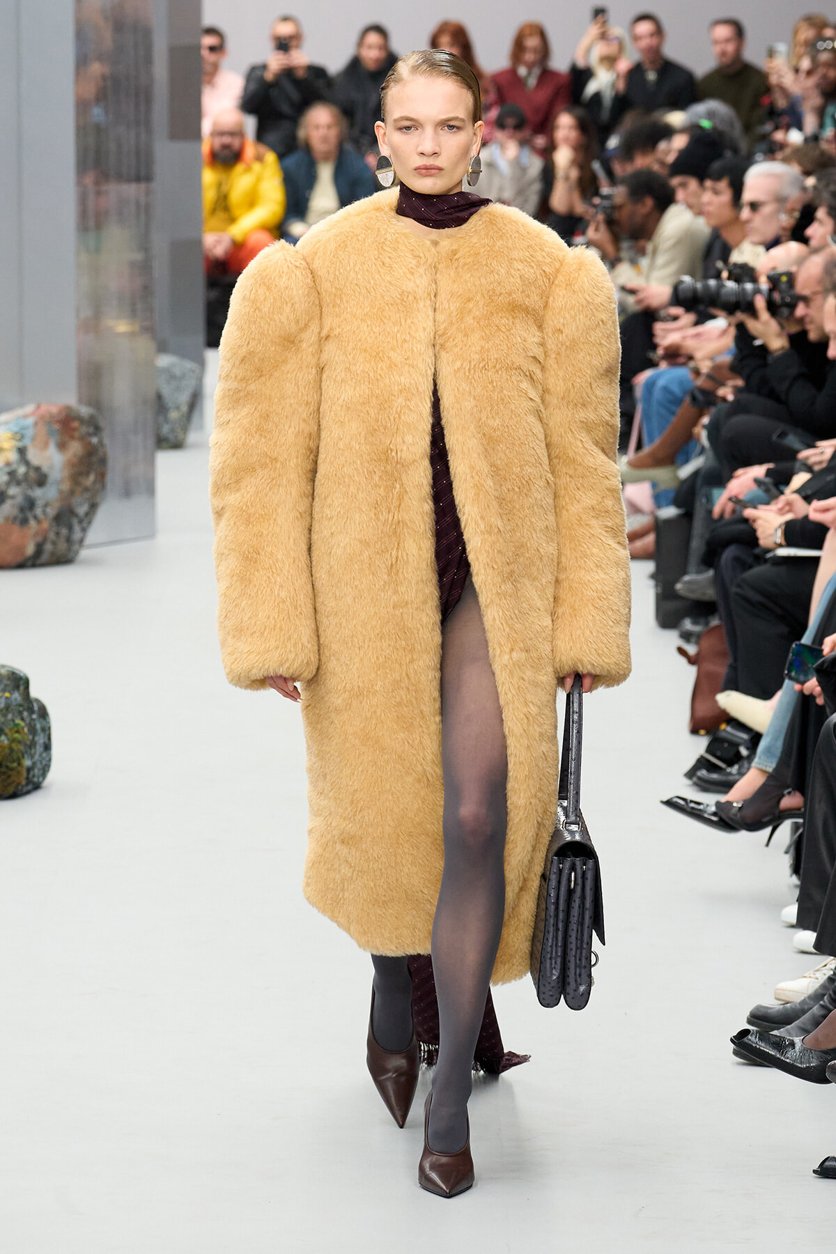 Runway model in a long tan fur coat over a burgundy outfit, carrying a black handbag as she walks the catwalk.