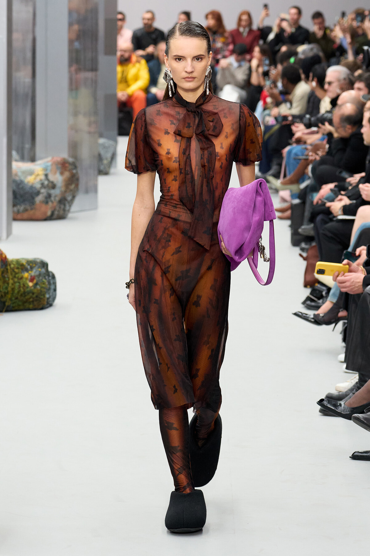 Model walks a runway in a sheer brown dress with a bow, holding a purple handbag; audience watches in the background.