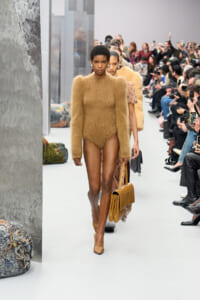 Model on a fashion runway wears a tan fuzzy bodysuit and tan heels, carrying a matching handbag as spectators watch.