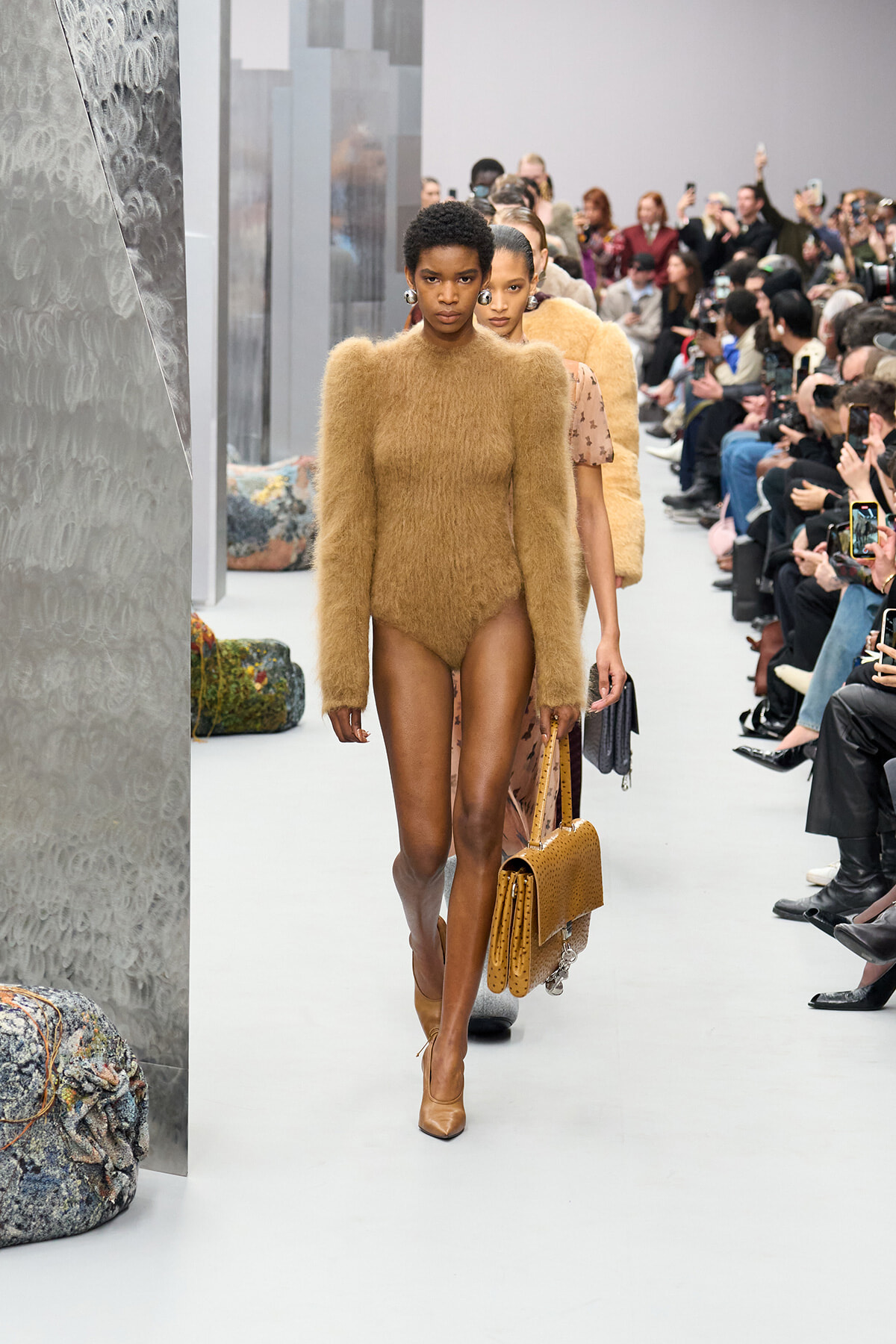 Model on a fashion runway wears a tan fuzzy bodysuit and tan heels, carrying a matching handbag as spectators watch.